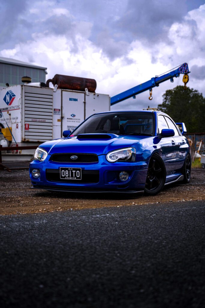 Lightning blue Subaru parked beside production plant retouched with Photoshop dodge & burn, gradient maps, and tonal adjustments for cinematic realism