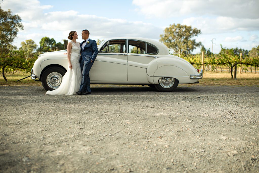 Just-married couple standing beside vintage car, retouched with Photoshop dodge & burn, frequency separation, and golden hour LUTs for cinematic warmth and editorial elegance.