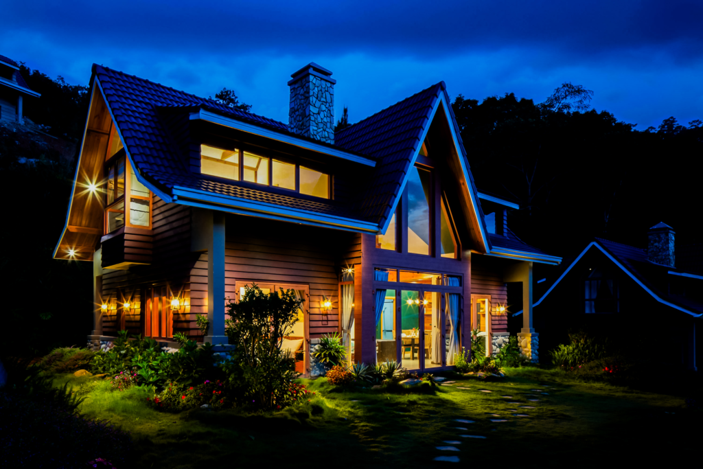 Timber cabin at dusk with illuminated windows and outdoor lights, showing cozy interior and landscaped approach.