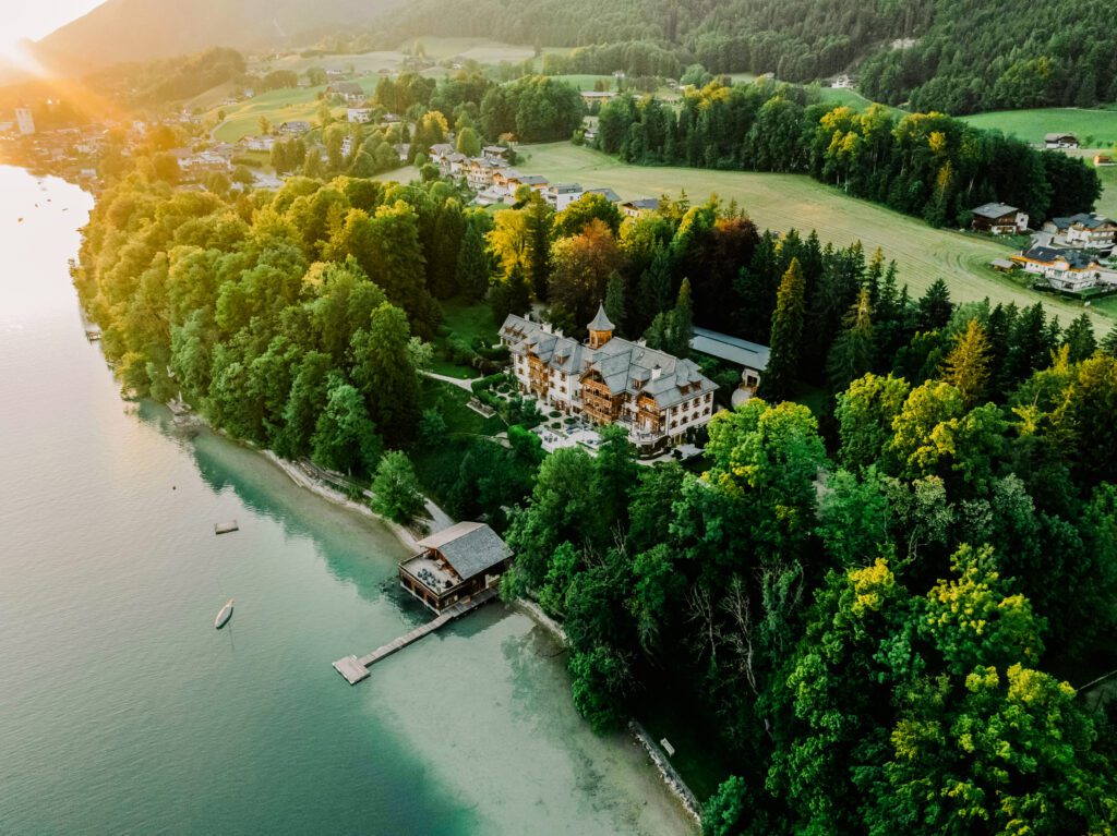 Dawn view of a castle-style residence nestled in trees with a lake house nearby, color-graded by la noir’e Da Vinci.